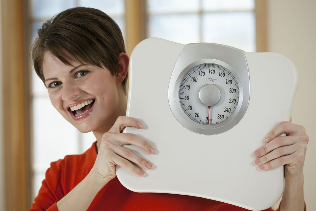 Attractive young woman holds a bathroom scale up while smiling at the camera. Horizontal shot