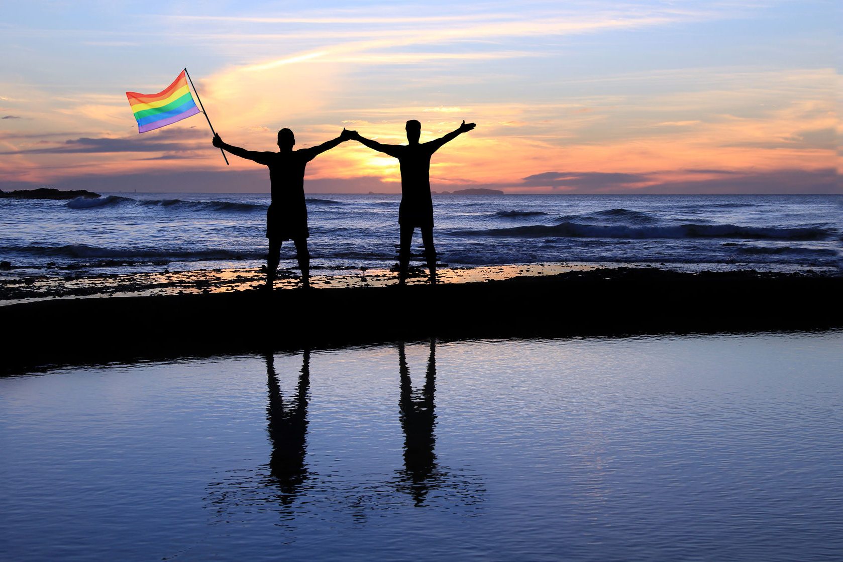 37337529 - silhouette of a gay couple holding a rainbow pride flag at sunset.